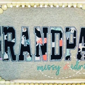May include: A gray t-shirt with the word "GRANDPA" in large, bold, black letters with a patterned fill. The text "missy & idris" is embroidered below in teal and coral. The shirt is decorated with white beads.
