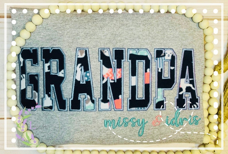 May include: A gray t-shirt with the word "GRANDPA" in large, bold, black letters with a patterned fill. The text "missy & idris" is embroidered below in teal and coral. The shirt is decorated with white beads.