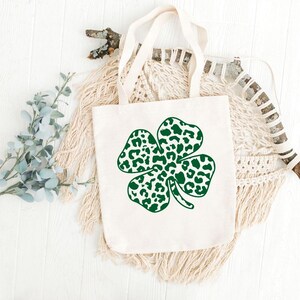 Pode incluir: Uma bolsa tote branca com um design de trevo de quatro folhas verde. O trevo &eacute; feito de manchas de estampa de leopardo.
