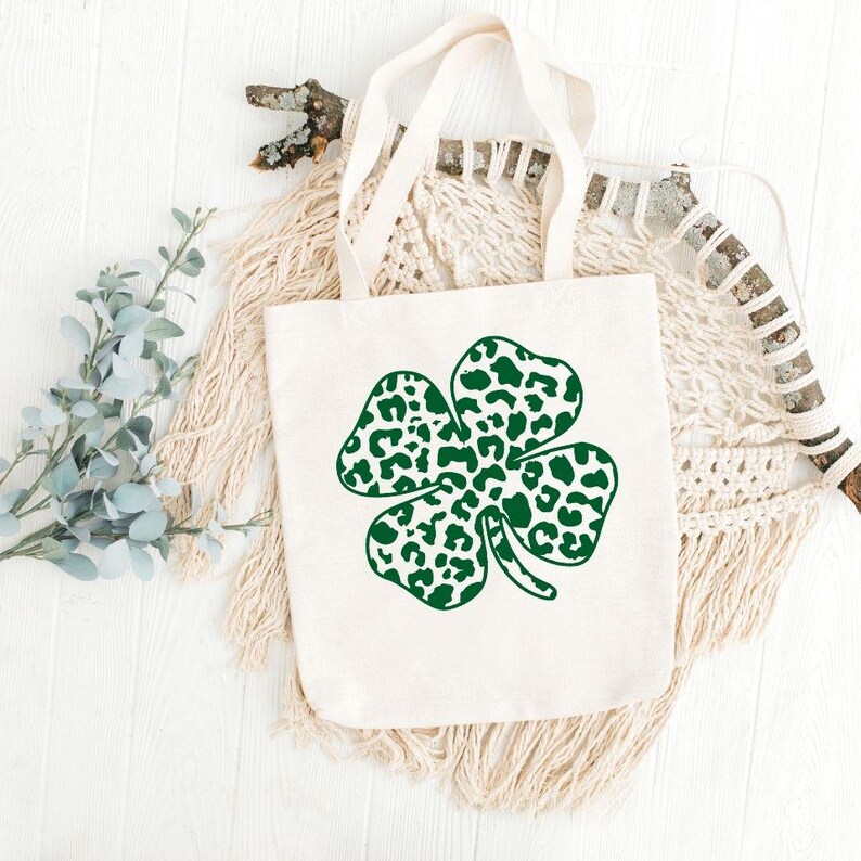 Pode incluir: Uma bolsa tote branca com um design de trevo de quatro folhas verde. O trevo &eacute; feito de manchas de estampa de leopardo.