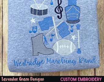 Puede incluir: Sudadera azul claro con bordados de banda de música. Presenta un tambor, notas musicales, una clave de sol, un uniforme de banda, banderas, un balón de fútbol americano y una bota de banda de música. El texto "Westridge Marching Band" está bordado en azul.