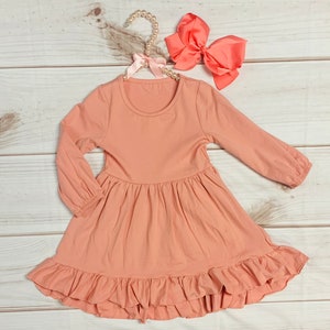May include: A peach-colored long-sleeved dress with a ruffled hem. The dress has a round neckline and is made of a soft, stretchy fabric.