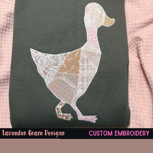 Pode incluir: Camisola verde escuro com um desenho de pato patchwork bordado em bege, castanho e rosa. O pato é feito de vários padrões de tecido. O texto "Lavender Grace Designs" e "CUSTOM EMBROIDERY" está na parte inferior.