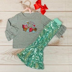 May include: A grey long-sleeve shirt with a ruffled sleeve and a pumpkin graphic. The shirt is paired with a teal sequin skirt.