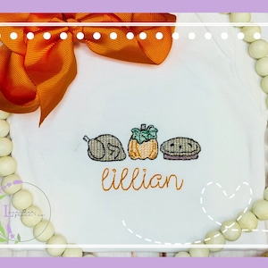 Può includere: Un body bianco con immagini ricamate a tema del Ringraziamento di un tacchino, una zucca e una torta. Il body è personalizzato con il nome "Lillian".