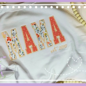 May include: White sweatshirt with the word "MAMA" in large, embroidered letters. The letters are coral with white polka dots and floral patterns. The text "est 2010" is embroidered below the word "MAMA".