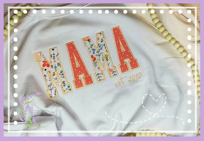 May include: White sweatshirt with the word "MAMA" in large, embroidered letters. The letters are coral with white polka dots and floral patterns. The text "est 2010" is embroidered below the word "MAMA".