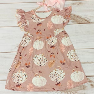 Puede incluir: Un vestido rosa sin mangas con estampado de calabazas blancas y marrones. El vestido tiene un cuello y mangas con volantes.