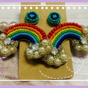 May include: A pair of colorful beaded rainbow earrings. The earrings feature a rainbow design with red, orange, yellow, green, and blue beads. The rainbow is attached to a cloud-shaped base with faux pearls and clear crystals. The earrings have a post backing.