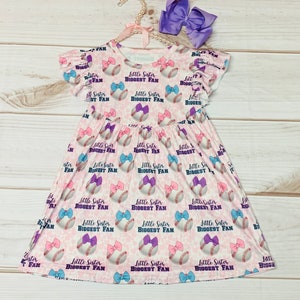 Puede incluir: Un vestido rosa y blanco con estampado de béisbol. El vestido tiene un volante en las mangas y dice "Little Sister Biggest Fan" en la parte delantera.