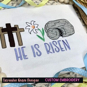 May include: White fabric with embroidered Easter theme. Features three crosses, a white lily, and a gray stone tomb. The text "HE IS RISEN" is in blue. The design includes the text "Lavender Grace Designs" and "CUSTOM EMBROIDERY".