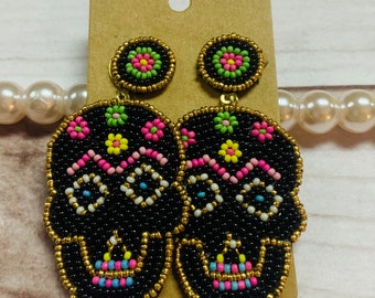 May include: Black beaded skull earrings with gold trim and post. The earrings feature colorful floral accents in pink, green, and yellow. The earrings are attached to a brown card.