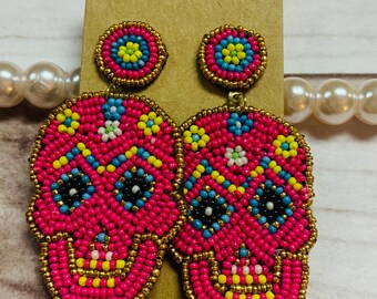 May include: A pair of pink beaded skull earrings with colorful accents. The earrings are dangle style and have a small round bead at the top.