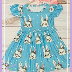 May include: A light blue dress with a white polka dot pattern and a print of bunnies wearing glasses. The dress has short, ruffled sleeves and a gathered skirt. The dress is on a hanger with a pink bow.