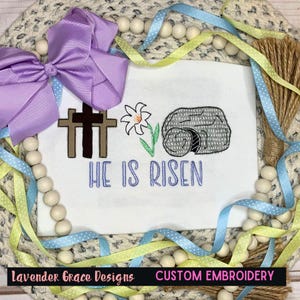 May include: White fabric with embroidered crosses, a flower, and a tomb with the text "HE IS RISEN" in blue. A purple bow and ribbons in blue and yellow surround the fabric. The text "Lavender Grace Designs CUSTOM EMBROIDERY" is at the bottom.