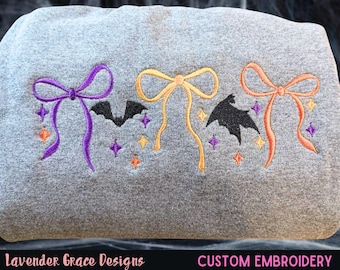 May include: A grey sweatshirt featuring embroidered Halloween designs: bows, bats, and stars in purple, orange, and yellow. The text "Lavender Grace Designs" and "CUSTOM EMBROIDERY" are at the bottom.