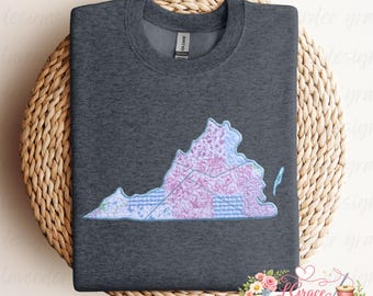 Könnte beinhalten: Dunkelgraues Sweatshirt mit einer gestickten Karte von Virginia in Rosa-, Blau- und Grüntönen. Das Kartendesign enthält florale und geometrische Muster. Das Sweatshirt wird auf einer gewebten Oberfläche präsentiert, mit einem floralen Logo unten rechts.