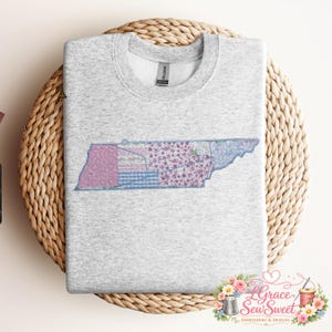 Puede incluir: Una sudadera gris claro con un diseño de patchwork bordado del estado de Tennessee. El diseño incorpora varios patrones, incluyendo florales, cuadros y texturas acolchadas, en tonos de rosa, morado y azul. La sudadera se muestra sobre una superficie de mimbre tejido.