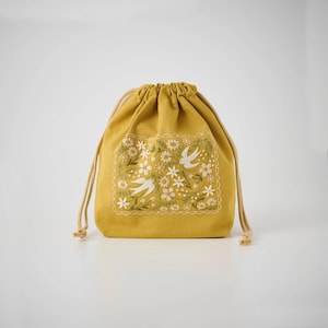 May include: A mustard yellow drawstring pouch featuring a rectangular embroidered panel on the front. The embroidery depicts white birds and various small flowers in shades of white and green, framed by a scalloped stitched border.