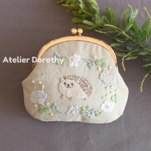 Könnte beinhalten: Eine hellgrüne, bestickte Geldbörse mit goldenem Verschluss. Die Geldbörse zeigt ein detailliertes Igel- und Blumendesign. Der Text "Atelier Dorothy" ist auf der linken Seite der Geldbörse zu sehen.