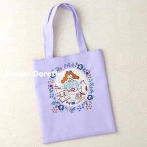 May include: A lavender tote bag with embroidered artwork. The artwork features a girl sewing, surrounded by a floral wreath, a cat, and sewing supplies. The text "Atelier Dorothy" is printed on the bag.