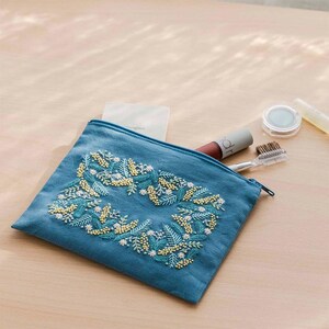May include: A teal blue zippered pouch with embroidered floral design in white, yellow, and teal. The pouch contains makeup items, including a lipstick, eyeshadow, and a brow brush. The pouch is on a light wood surface.
