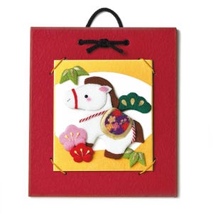 May include: A red square wall hanging with a decorative felt horse. The horse is white with a brown mane and tail, and a colorful saddle. The background is yellow and white, with floral and leaf accents. A black cord is attached for hanging.