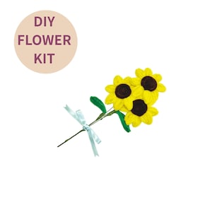 May include: A DIY flower kit featuring a bouquet of three crocheted sunflowers. The sunflowers have yellow petals and dark brown centers. Green stems and leaves support the flowers, tied with a light blue ribbon. The kit includes the text "DIY FLOWER KIT".