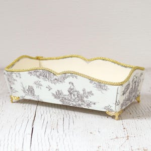 Könnte beinhalten: Rechteckige dekorative Box mit gewelltem Rand, mit einem Toile-Muster in Grau und Weiß. Die Box hat goldfarbene Verzierungen und kleine goldfarbene Füße, die einen Hauch von Eleganz verleihen. Der Innenraum ist cremefarben.
