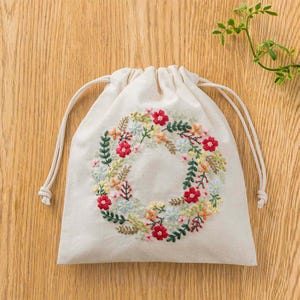 May include: A cream-colored drawstring bag with an embroidered floral wreath design. The wreath features red, blue, yellow, and green flowers and leaves. The bag is made of a natural fabric and has a simple, elegant design.