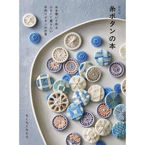 Puede incluir: Una colección de botones de hilo hechos a mano en varios diseños y colores, incluyendo blanco, azul y beige, dispuestos en un plato gris. Los botones presentan intrincados patrones y texturas tejidas. Texto japonés visible.