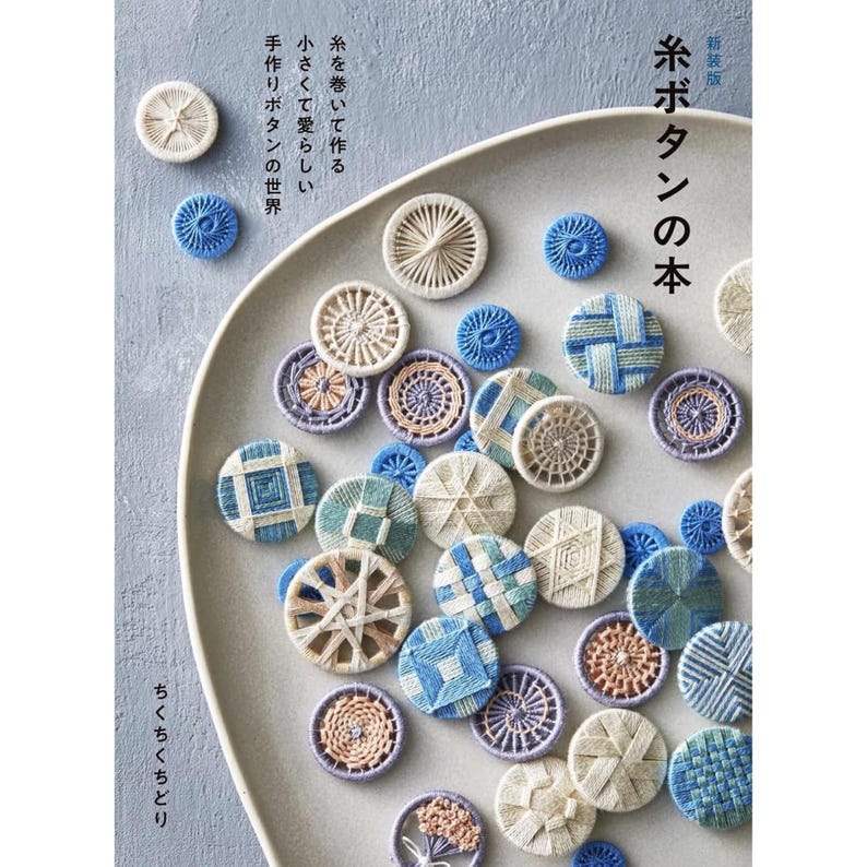 Puede incluir: Una colecci&oacute;n de botones de hilo hechos a mano en varios dise&ntilde;os y colores, incluyendo blanco, azul y beige, dispuestos en un plato gris. Los botones presentan intrincados patrones y texturas tejidas. Texto japon&eacute;s visible.