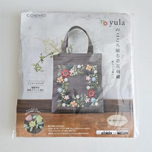 May include: A gray tote bag with a floral embroidery design. The bag features a square floral wreath with red, pink, yellow, and blue flowers. The bag has a handle and is packaged in a clear plastic bag. The text "yula" is visible.