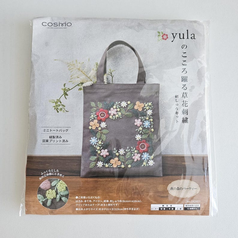 May include: A gray tote bag with a floral embroidery design. The bag features a square floral wreath with red, pink, yellow, and blue flowers. The bag has a handle and is packaged in a clear plastic bag. The text "yula" is visible.