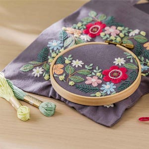 May include: A floral embroidery project in progress. A wooden embroidery hoop holds a gray fabric with a colorful embroidered flower design. Green, pink, and white flowers are featured. Skeins of green and yellow embroidery floss are on the wooden surface.