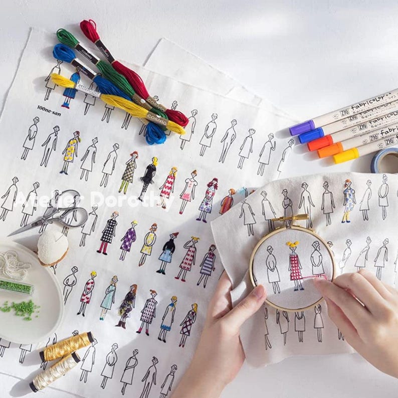 May include: White fabric with a repeating line drawing of figures, some of which are embroidered with colorful thread. Embroidery floss in red, green, blue, and yellow are visible. The image also shows a wooden embroidery hoop and colored markers.