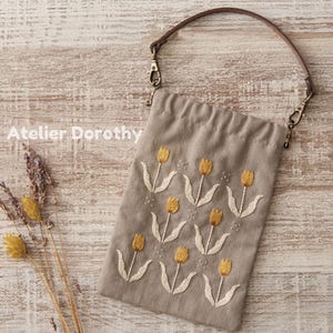 May include: A beige drawstring bag with a brown leather handle and gold-colored clasps. The bag is embroidered with yellow tulips and white stems. The text "Atelier Dorothy" is printed on the bag.