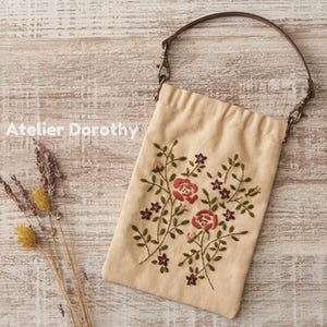 May include: Beige fabric bag with a floral embroidery design featuring red roses and purple flowers. The bag has a brown leather handle and metal clasps. The text "Atelier Dorothy" is visible on the left side.