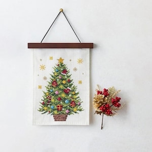 May include: A cross-stitch Christmas tree wall hanging with a wooden bar and black string for hanging. The tree is decorated with ornaments and a star. A sprig of red berries and gold pinecone is to the right.