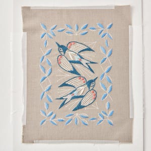 May include: A rectangular piece of beige fabric featuring an embroidered design of two birds in flight, colored in shades of blue, white, and coral. The birds are surrounded by a rectangular border of embroidered blue leaves on a light background.