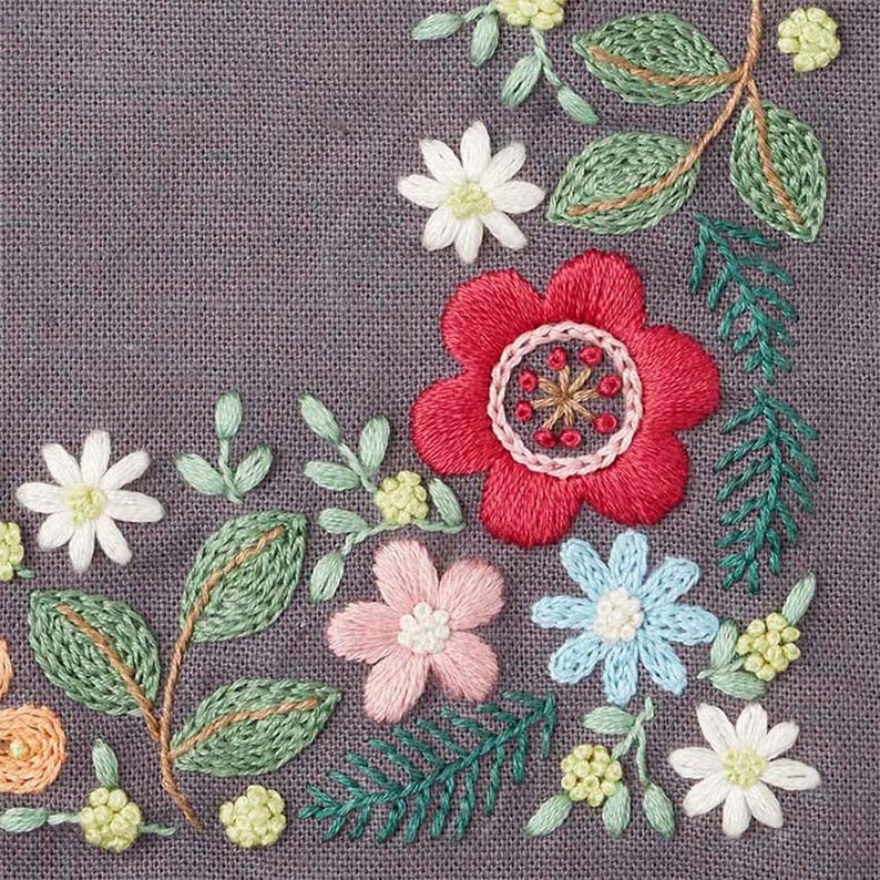 May include: Close-up of a floral embroidery design on gray fabric. The design features a large red flower with a detailed center, surrounded by smaller white, pink, and blue flowers, green leaves, and small yellow buds. The stitches are visible.