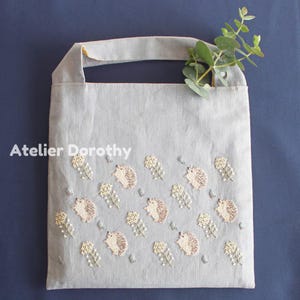Puede incluir: Un bolso tote gris claro con bordados de erizos y diseños florales. El bolso tiene una sola asa y el texto "Atelier Dorothy" está impreso en la parte delantera. Una ramita de vegetación está colocada en el bolso.
