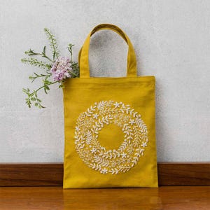 May include: Mustard yellow tote bag with white embroidered floral wreath design. The tote bag has two handles and is made of a sturdy fabric. A sprig of flowers is placed inside the bag.