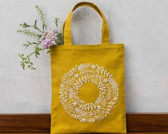 May include: Mustard yellow tote bag with white embroidered floral wreath design. The tote bag has two handles and is made of a sturdy fabric. A sprig of flowers is placed inside the bag.