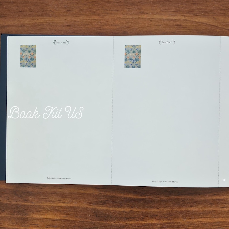 May include: Open book with the text "Book Kit US" in a cursive font. The book has two small floral design images and the words "Post Card" above them. The book is on a wooden surface.