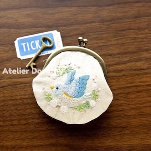 Puede incluir: Un pequeño monedero blanco roto con cierre metálico, con un pájaro azul bordado rodeado de hojas verdes y flores blancas. Una llave de estilo vintage y un ticket con la palabra "TICK" están junto al monedero.