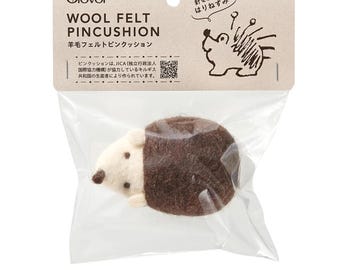 May include: A wool felt pincushion shaped like a hedgehog, packaged in a clear plastic bag. The pincushion is brown and cream-colored, with the text "WOOL FELT PINCUSHION" and Japanese text on the packaging. The Clover logo is visible.