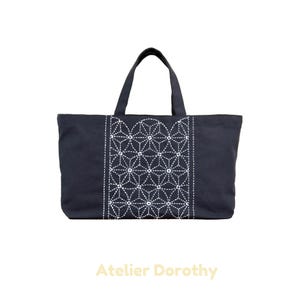 Può includere: Una borsa tote blu navy con ricami geometrici bianchi. La borsa ha una forma rettangolare con due manici e il testo "Atelier Dorothy" in basso.