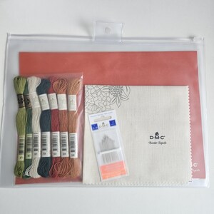 May include: A clear plastic zippered pouch containing embroidery supplies. The kit includes skeins of thread in various colors, a needle packet, a piece of fabric with a floral design, and a coral-colored paper. The DMC brand is visible.