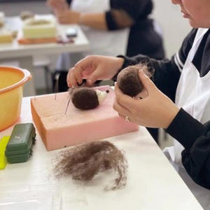 May include: A craft project in progress, featuring a small, brown and white felt hedgehog on a pink foam block. The image shows the process of needle felting, with loose brown wool and a ball of wool in the crafter's hands.